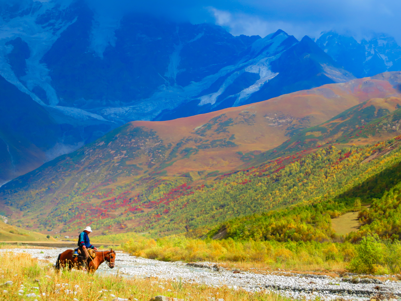 A man on a horse in the middle of the field by a river with a large glacier in the background