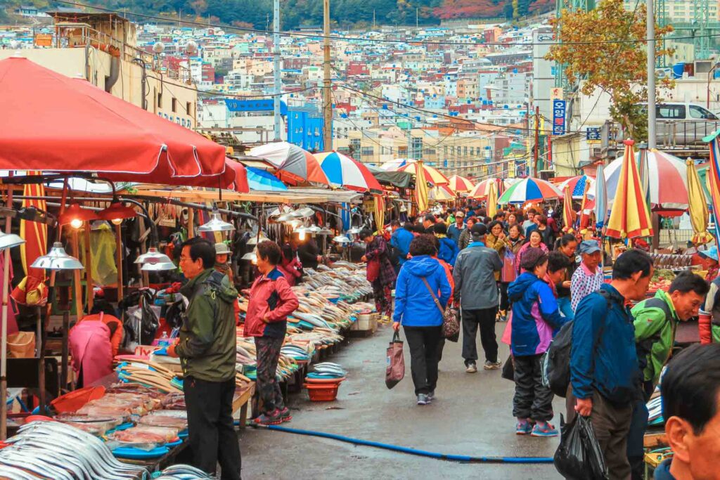 Many people and market stalls in the streets of Busan with many colourful houses in the background on top of the mountain