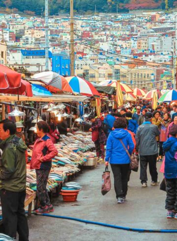 Many people and market stalls in the streets of Busan with many colourful houses in the background on top of the mountain