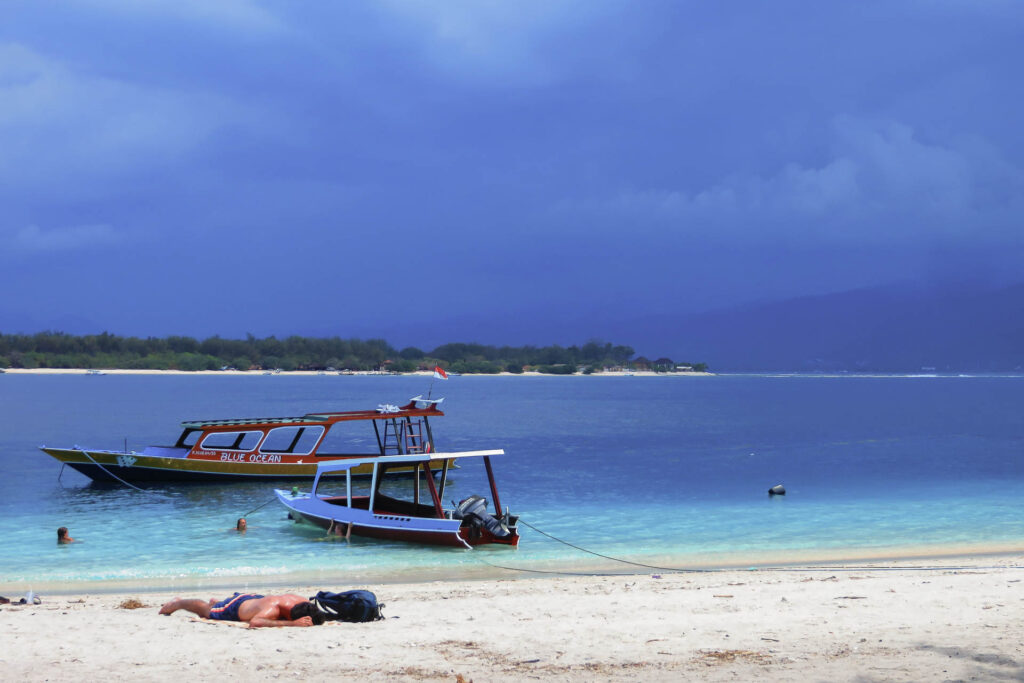 A man laying in the sand with a blue sky with a couple of boats moored in the Gili Islands
