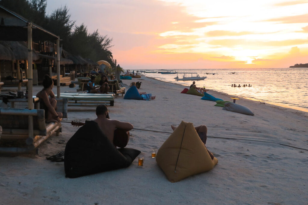 The sun setting in the horizon of the sea with people in the water and others sitting drinking. The front sand seats has two people, one playing guitar