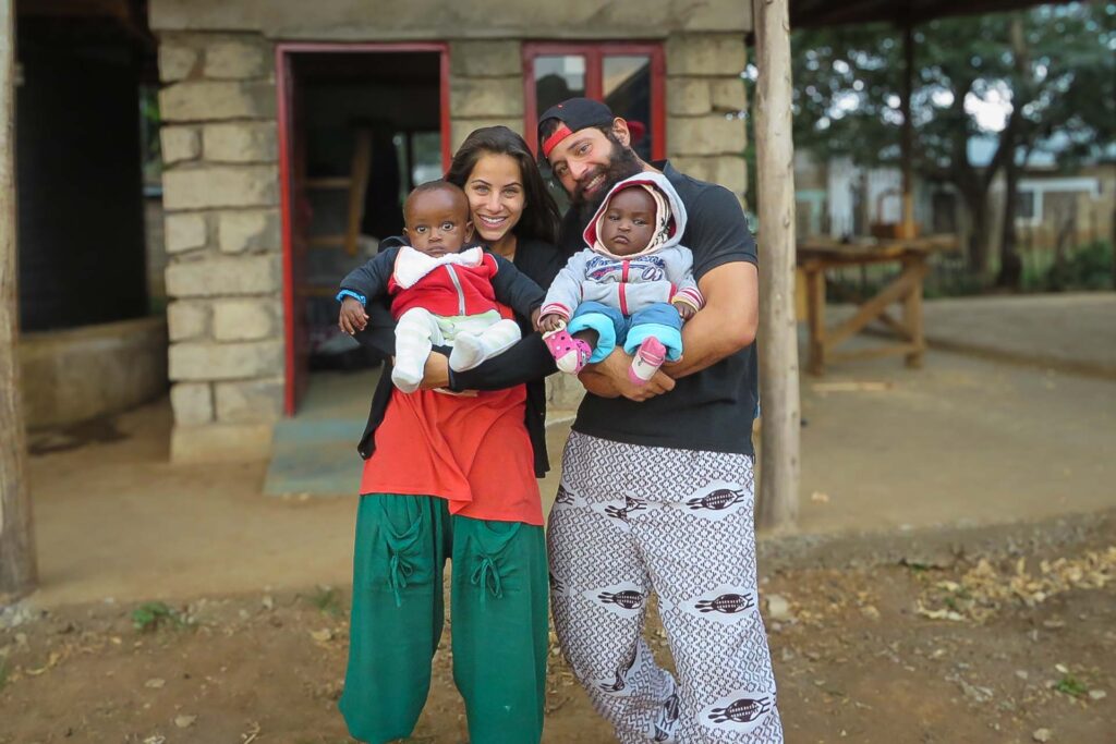 Tiago e Fernanda segurando um bebê de colo cada um em seus braços na frente de uma construção ainda não terminada no orfanato no Quênia