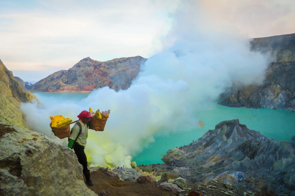 A man carrying sulphur in two baskets on his shoulder up the Ijen volcano with the acid lake in the background