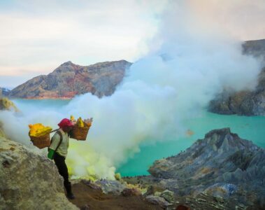 A man carrying sulphur in two baskets on his shoulder up the Ijen volcano with the acid lake in the background