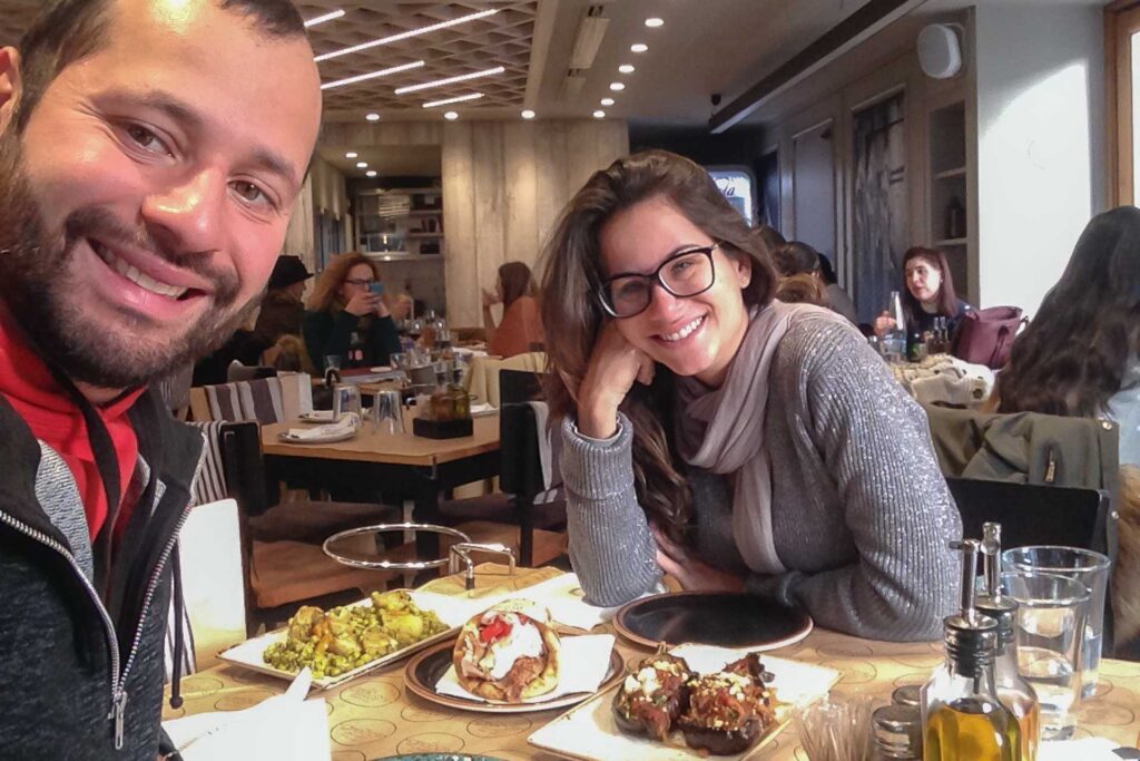 Fernanda e Tiago sentados no restaurante em Atenas comendo uma comida local