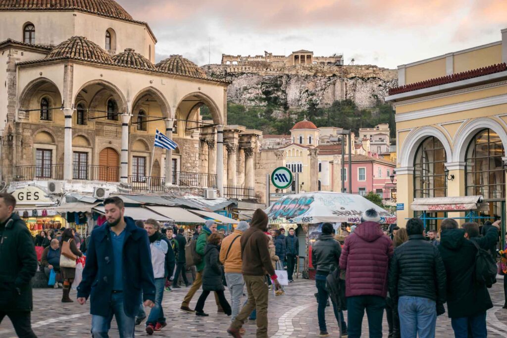 O bairro de Monastiraki na Grécia com uma praça cheia de pessoas andando e a Acrópoles no topo da montanha ao fundo