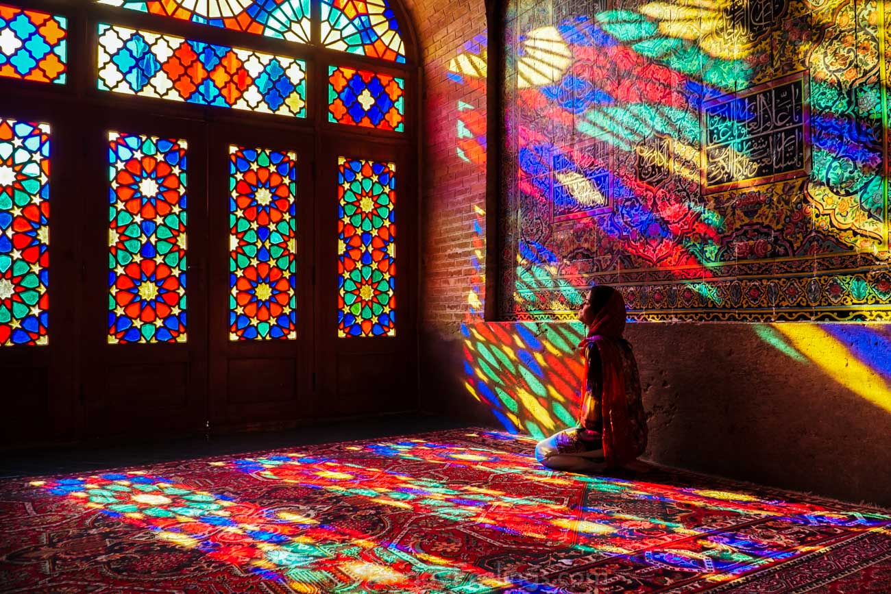 Fernanda de joelhos na Mesquita Rosa, com luzes dos vitrais todas coloridas iluminando ela, o carpete e a parede