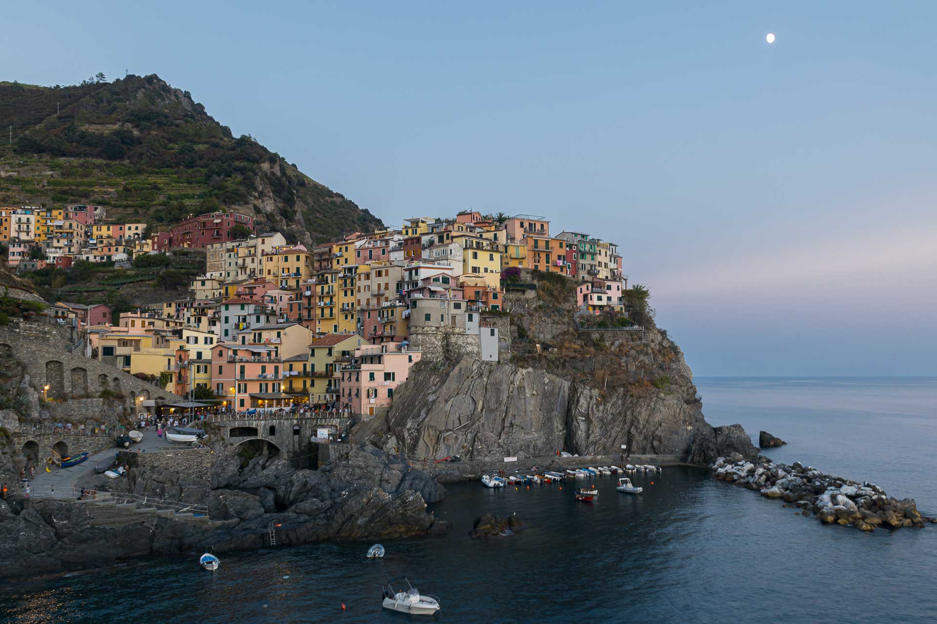 Manarola village of the Cinque Terre with some houses on top of a rock and others near the beach at twilight and the moon