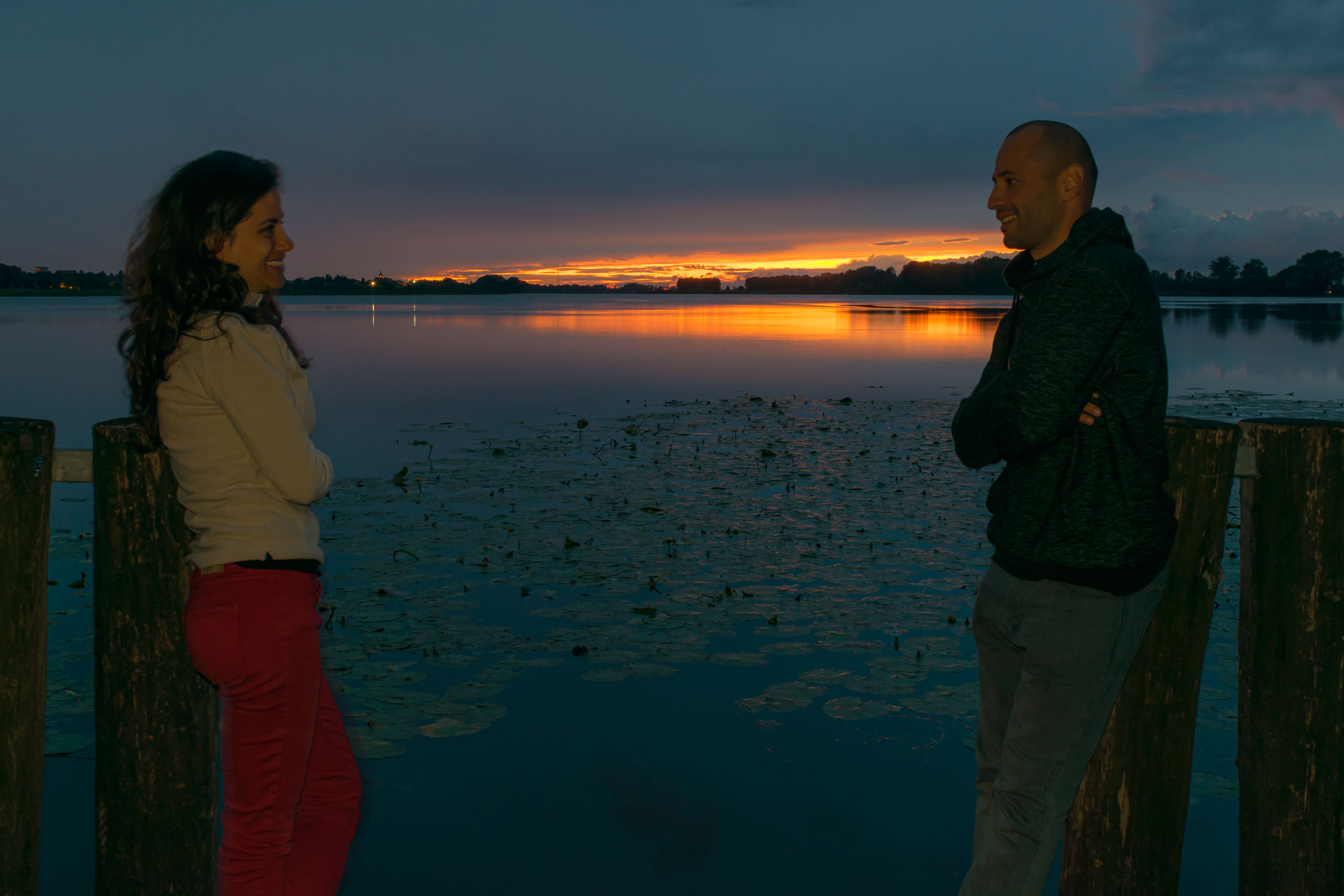 Tiago e Fernanda de frente um para o outro na frente do lago de Mantova na Italia enquanto o sol de poe ao fundo