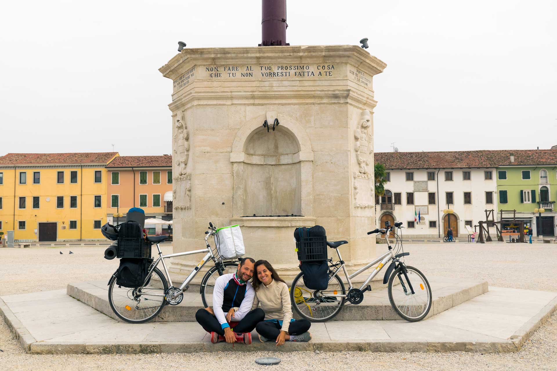 Tiago e Fernanda sentados na frente de um monumento com suas bibicletas em Palmonova, última cidade da viagem de bicicleta pela Europa