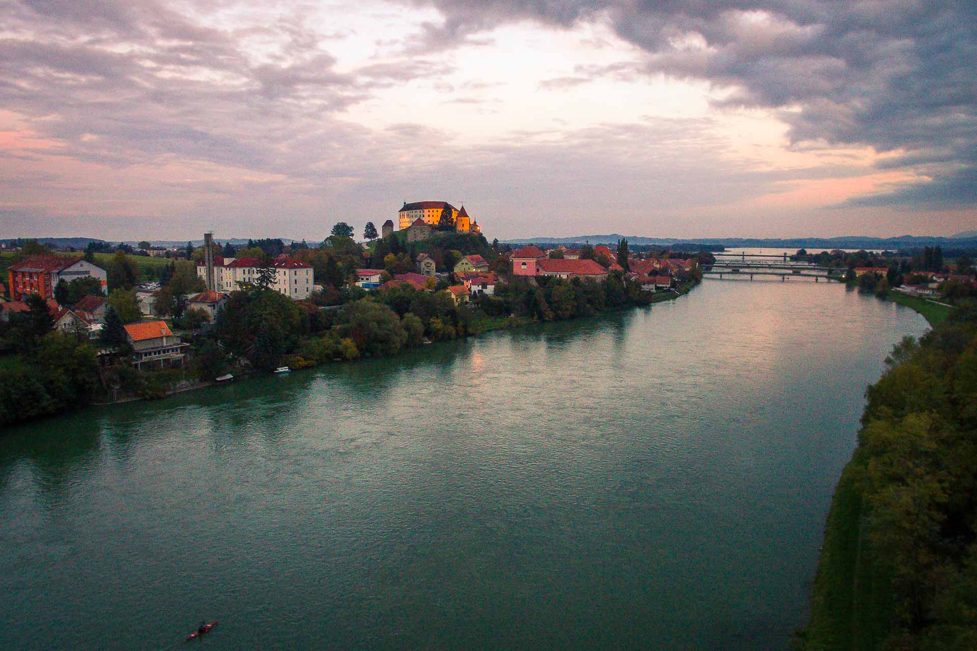A large river crossing the city with a castle on top of the mountain and a pinkish sky