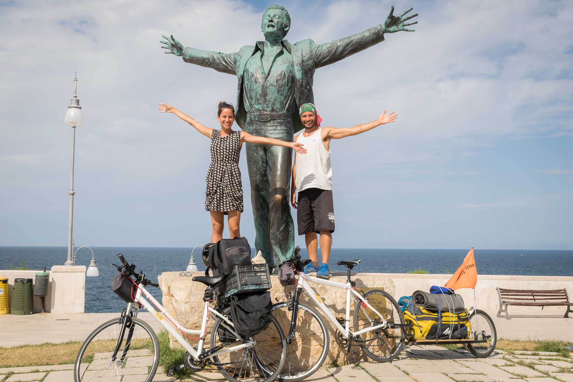 Tiago e Fernanda posando em frente a estátua de Domenico Modugno na Puglia com duas bicicletas em Polignano A Mare