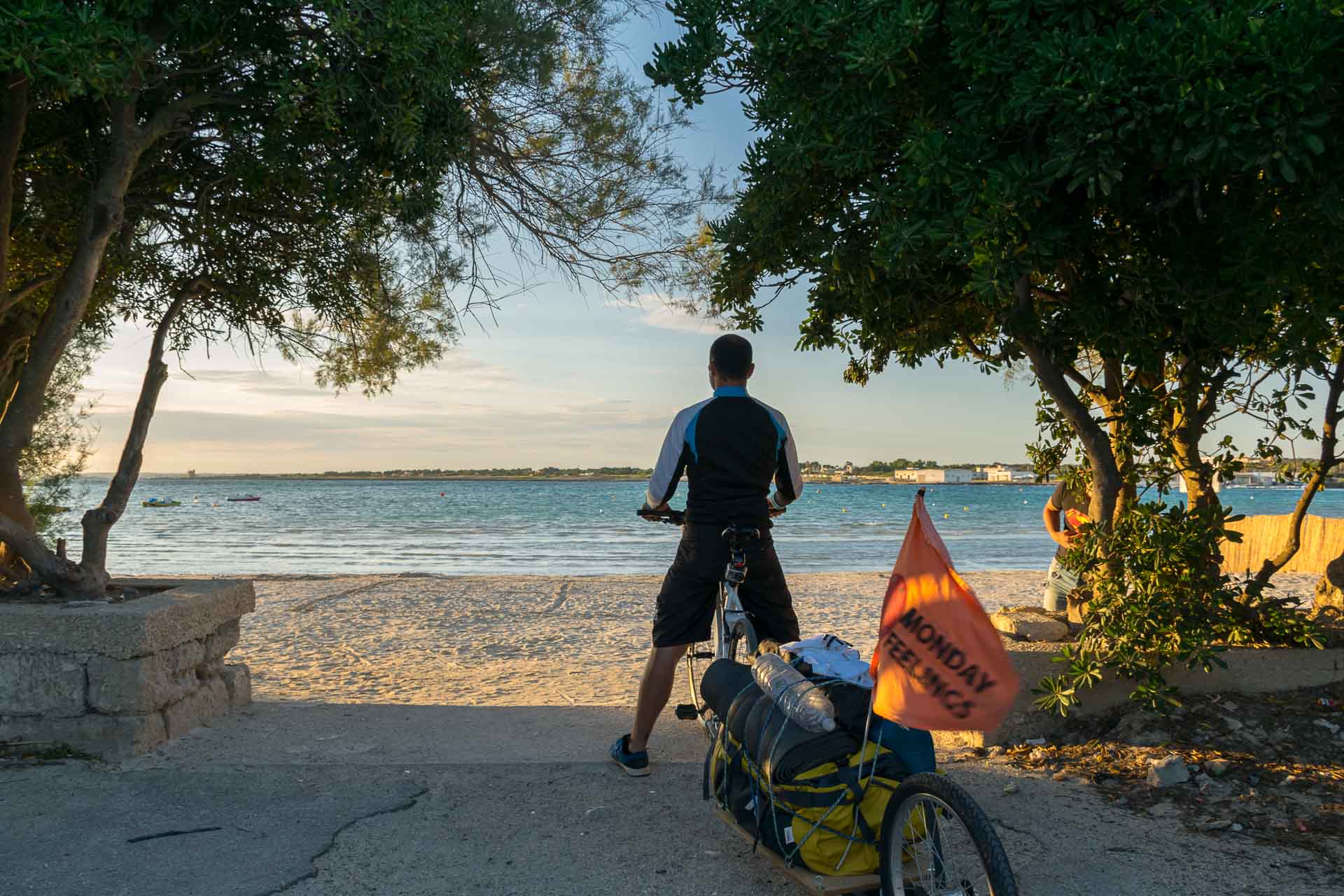 Tiago chegando de bicicleta na entrada da praia da Puglia entre duas arvores