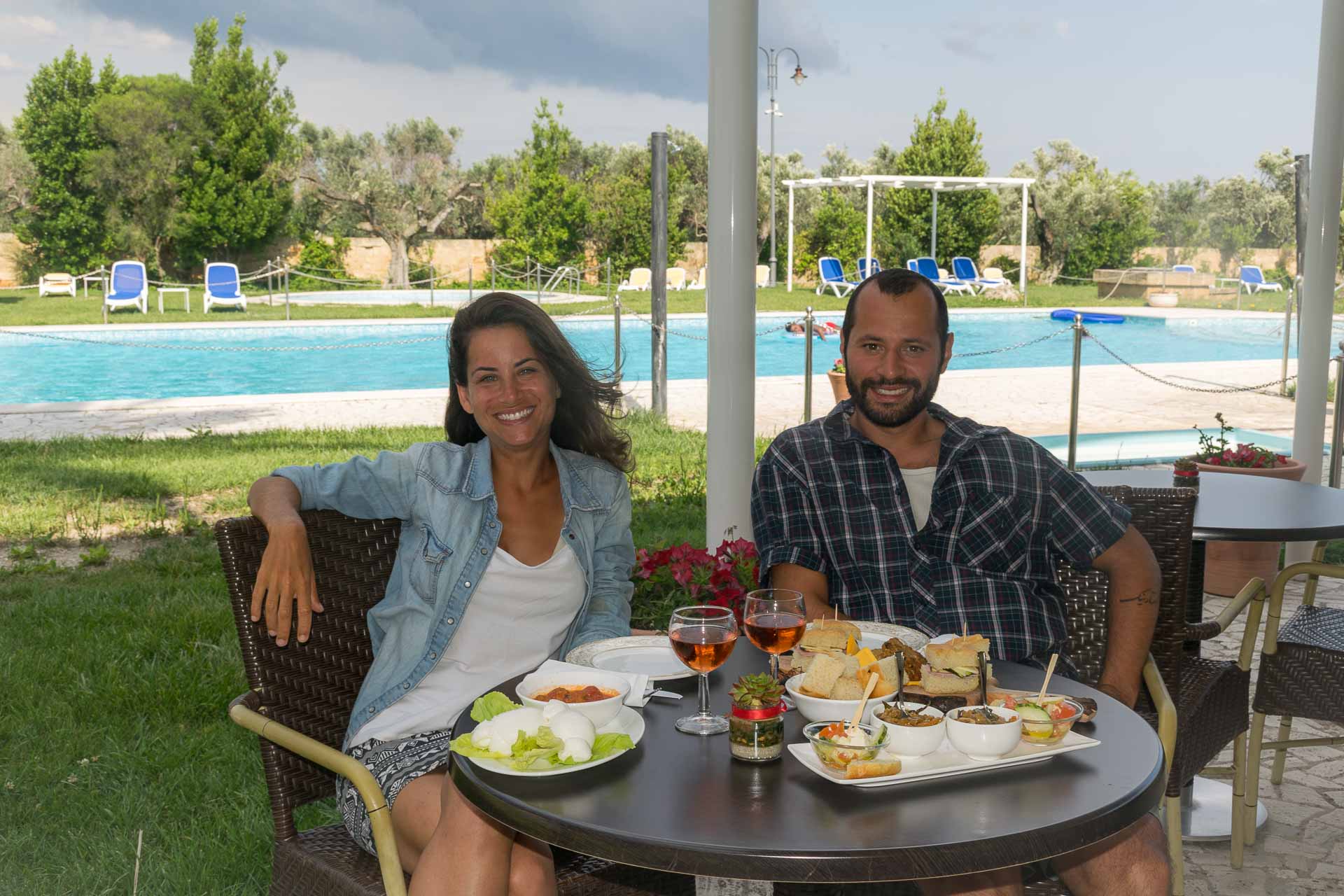 Tiago e Fernanda sentados na mesa da piscina bebendo vinho e comendo petiscos na masseria da Italia