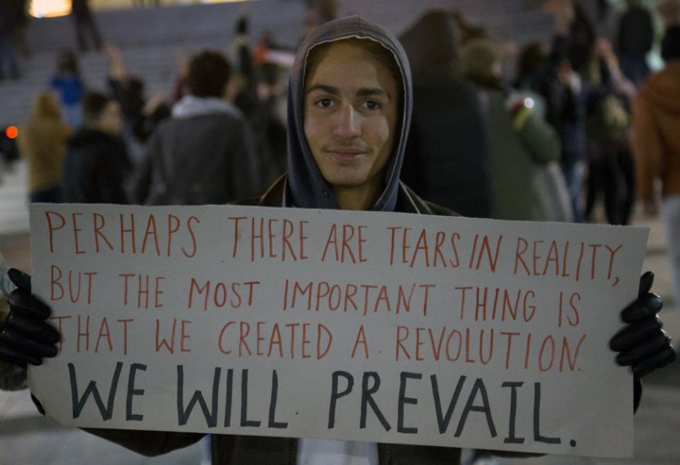 Um jovem Sírio segurando um cartaz dizendo: Talvez exista lágrimas na realidade, mas a coisa mais importante é que nós criamos uma revolução. Vamos prevalecer