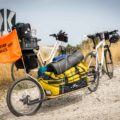 Two bikes alone next to each other with an attached trolley carrying a large yellow bag and a flag written Monday Feelings on top of a cliff overseeing the sea