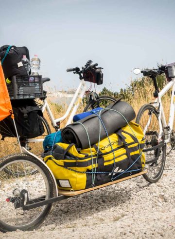 Two bikes alone next to each other with an attached trolley carrying a large yellow bag and a flag written Monday Feelings on top of a cliff overseeing the sea