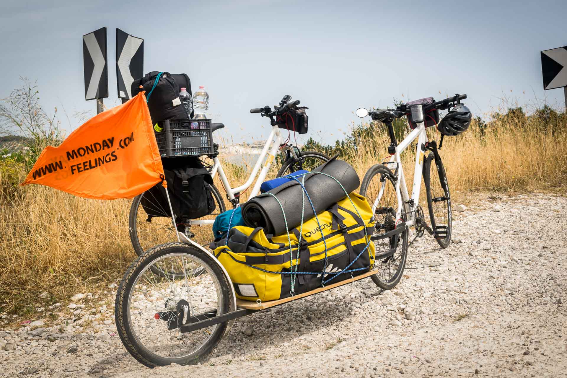 Two bikes alone next to each other with an attached trolley carrying a large yellow bag and a flag written Monday Feelings on top of a cliff overseeing the sea