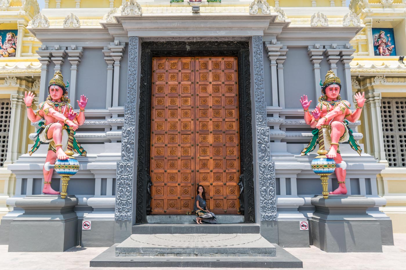 Fernanda sentada na frente da entrada de um templo de Penang com uma porta bem grande entre duas estátuas