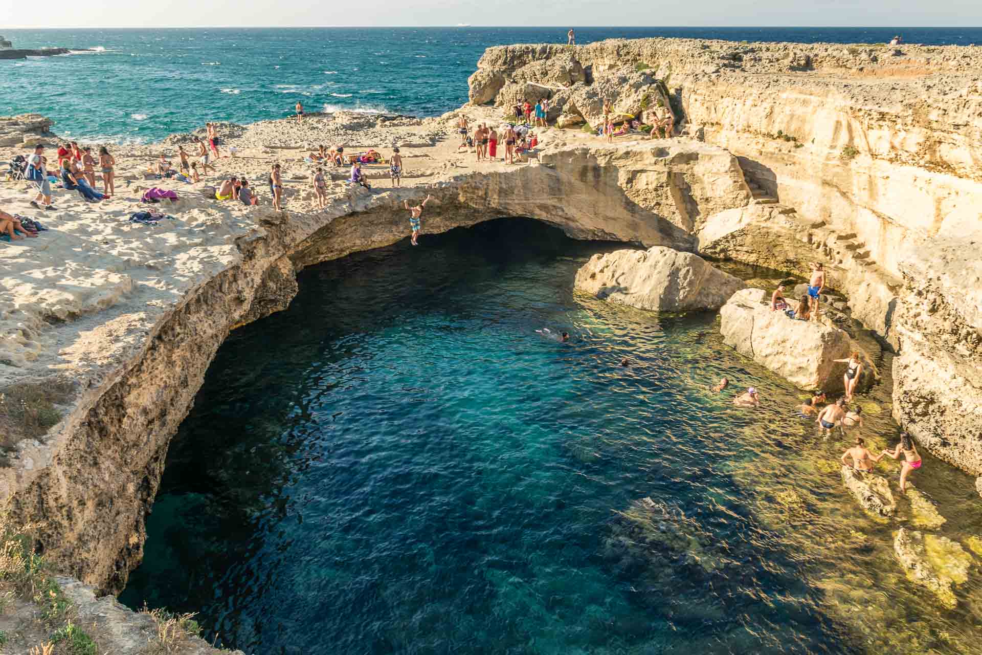 Uma grande piscina natural de água do mar no meio da rocha com o oceano ao fundo e muitas pessoas nadando e pulando