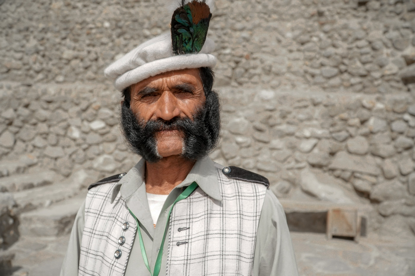 Um homem barbudo com vestimenta tradicional de Hunza e uma pena no chapeu