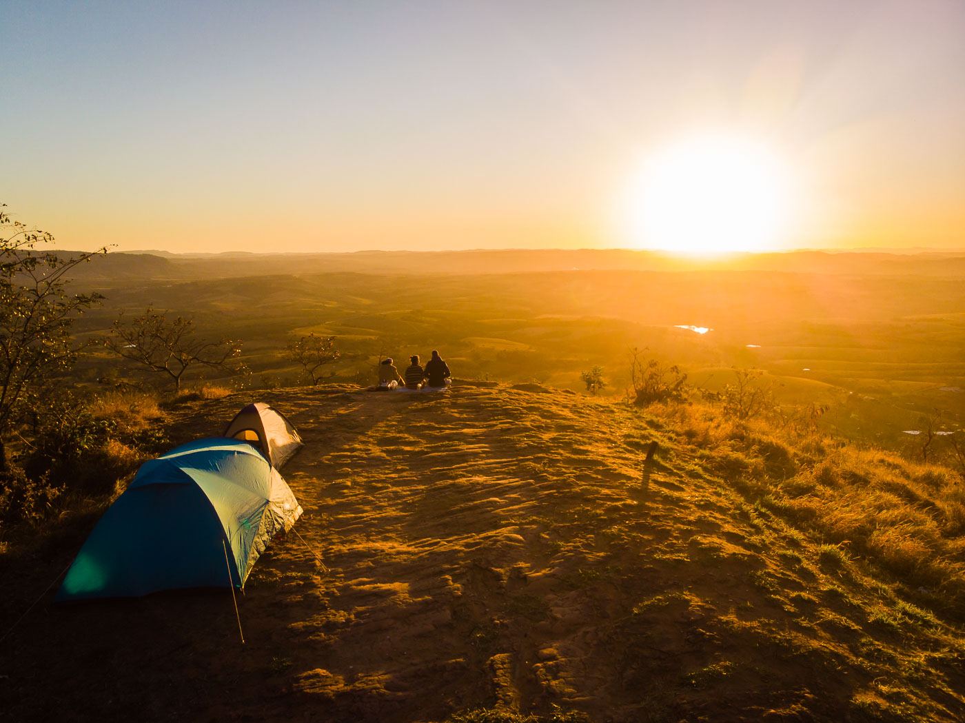 duas barracas no topo da montanha com três pessoas sentadas na beira da montanha vendo o nascer do sol no horizonte