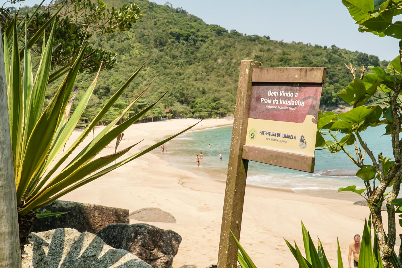 A welcome sign to the Indaiaúba Beach in Ilhabela and a large white sand beach by the mountain in the back