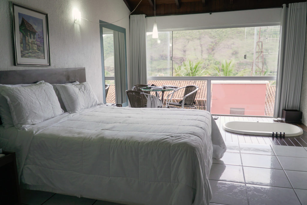 Quarto de casal no hotel Balneario do Lago em Capitólio com uma cama de casal, banheira e uma vista com um vidro gigante