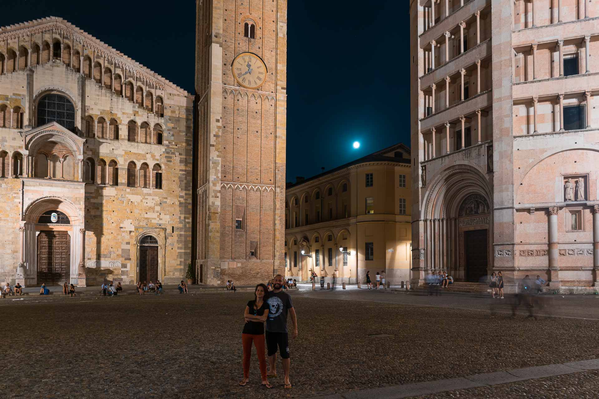 Vista da praça principal de Parma a noite com Tiago e Fernanda na frente do duome de Parma e do Batistério