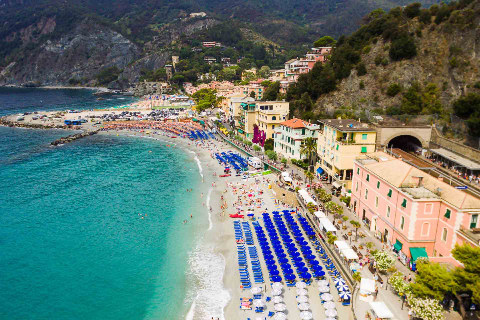 Vista aérea da praia de Cinque Terre cheia de guarda-sois e cadeiras com a estação de trem ao lado