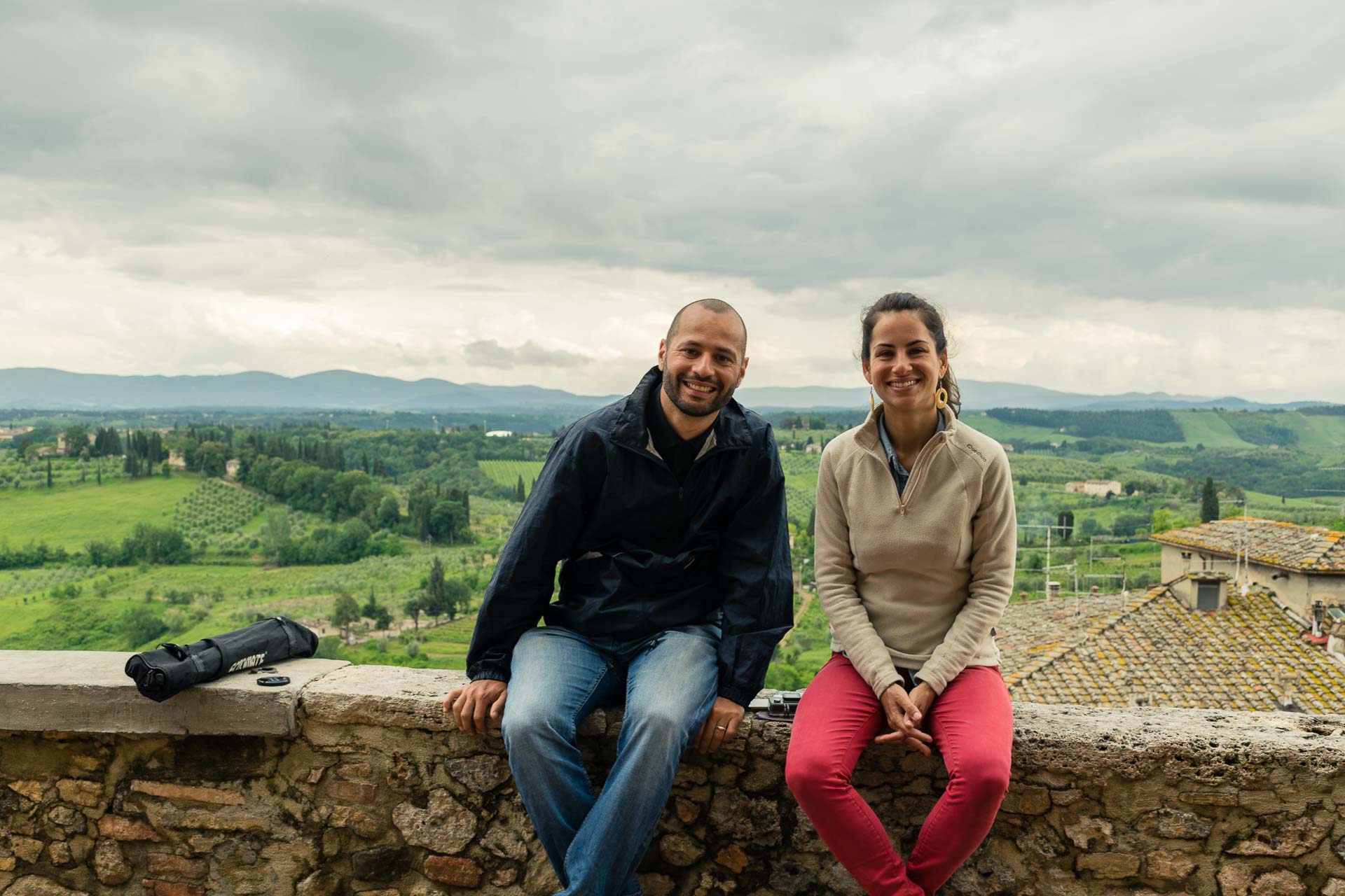 Tiago and Fernanda sitting in a wall with a large green field as background when cycling through the the best towns in Tuscany