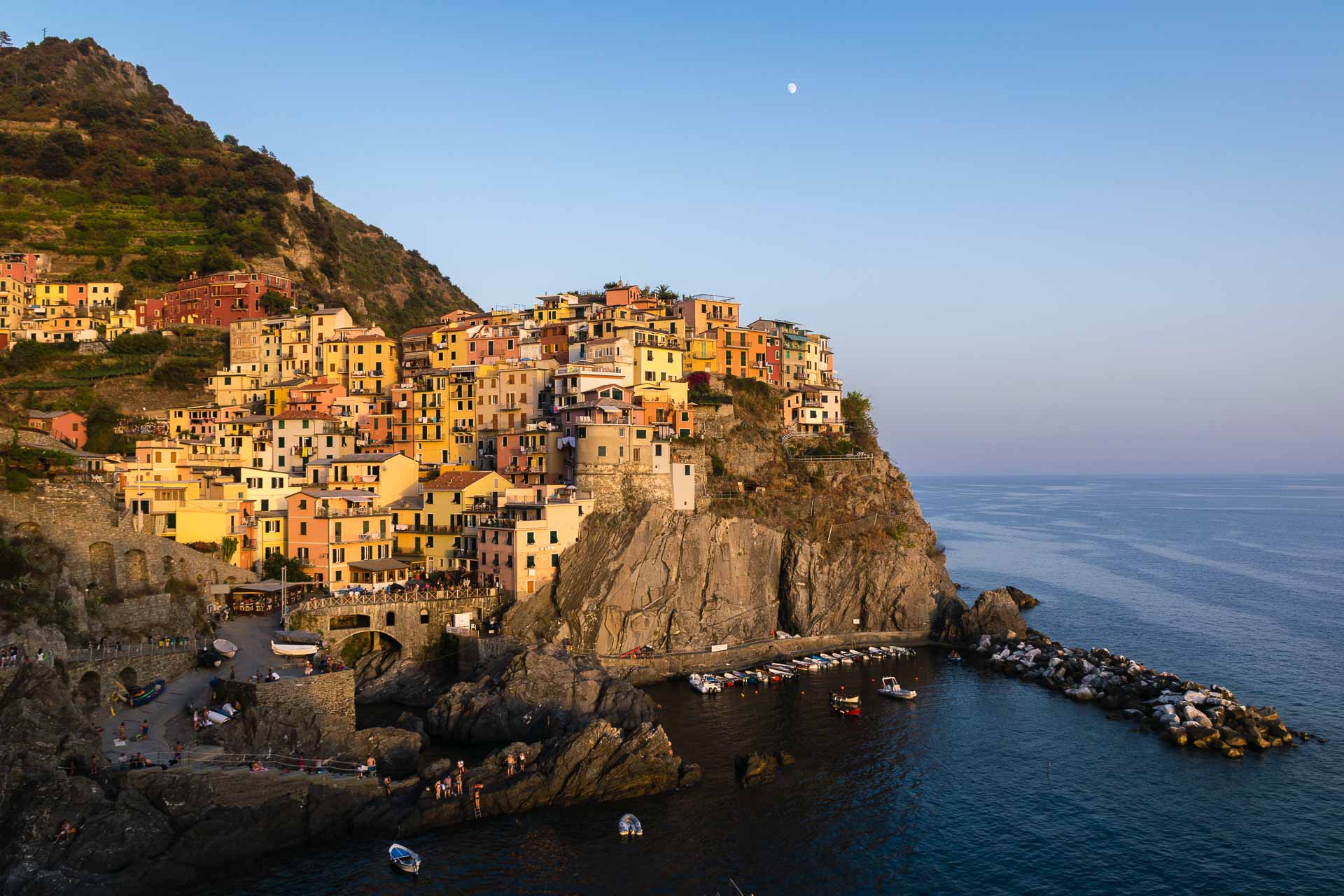 Uma das Cinque Terre iluminada pelo entardecer em cima da rocha na beira do mar com a lua no céu azul