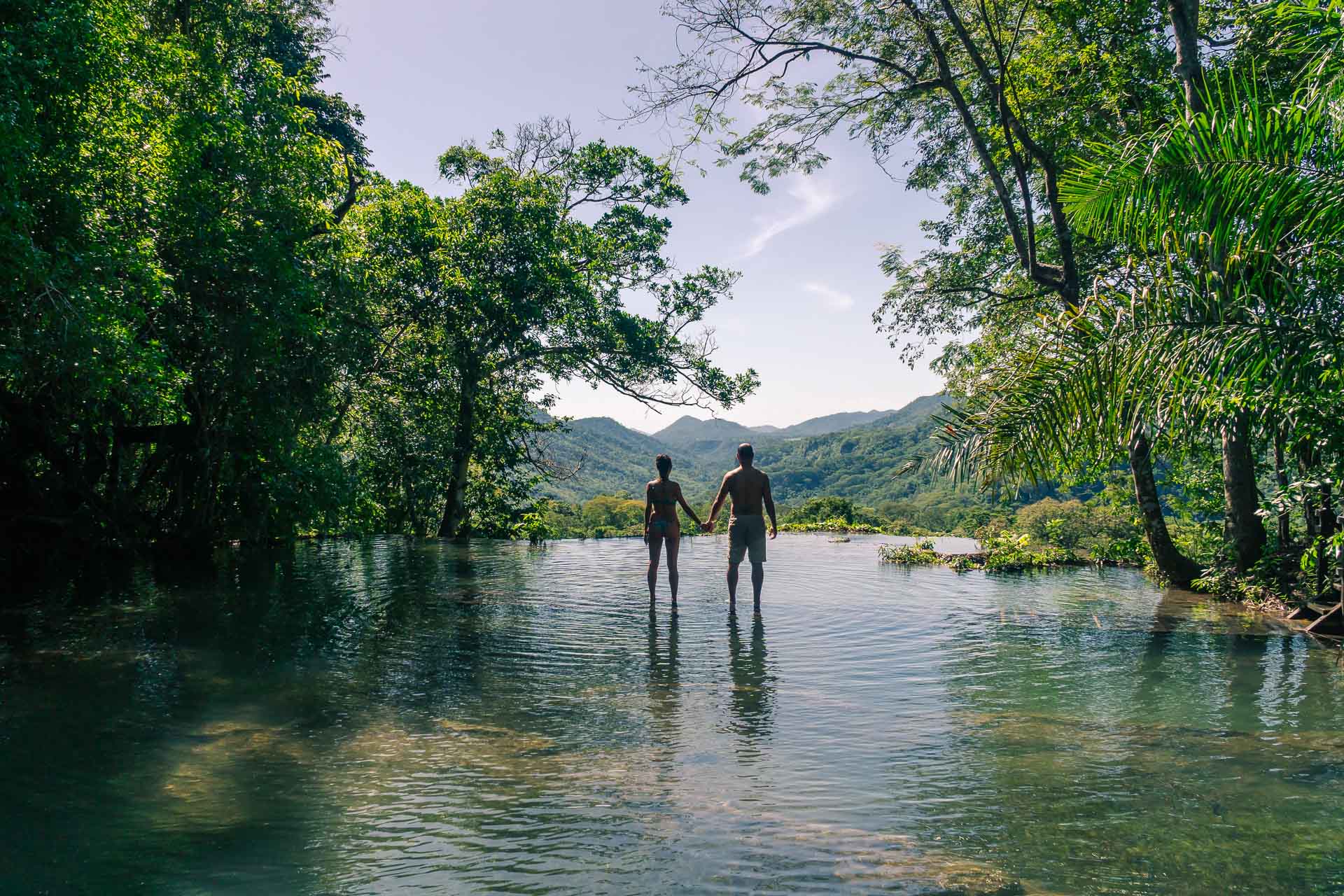 Tiago e Fernanda do Monday Feelings dando as mãos na beira de um lago com borda infinita vendo uma montanha e um penhasco