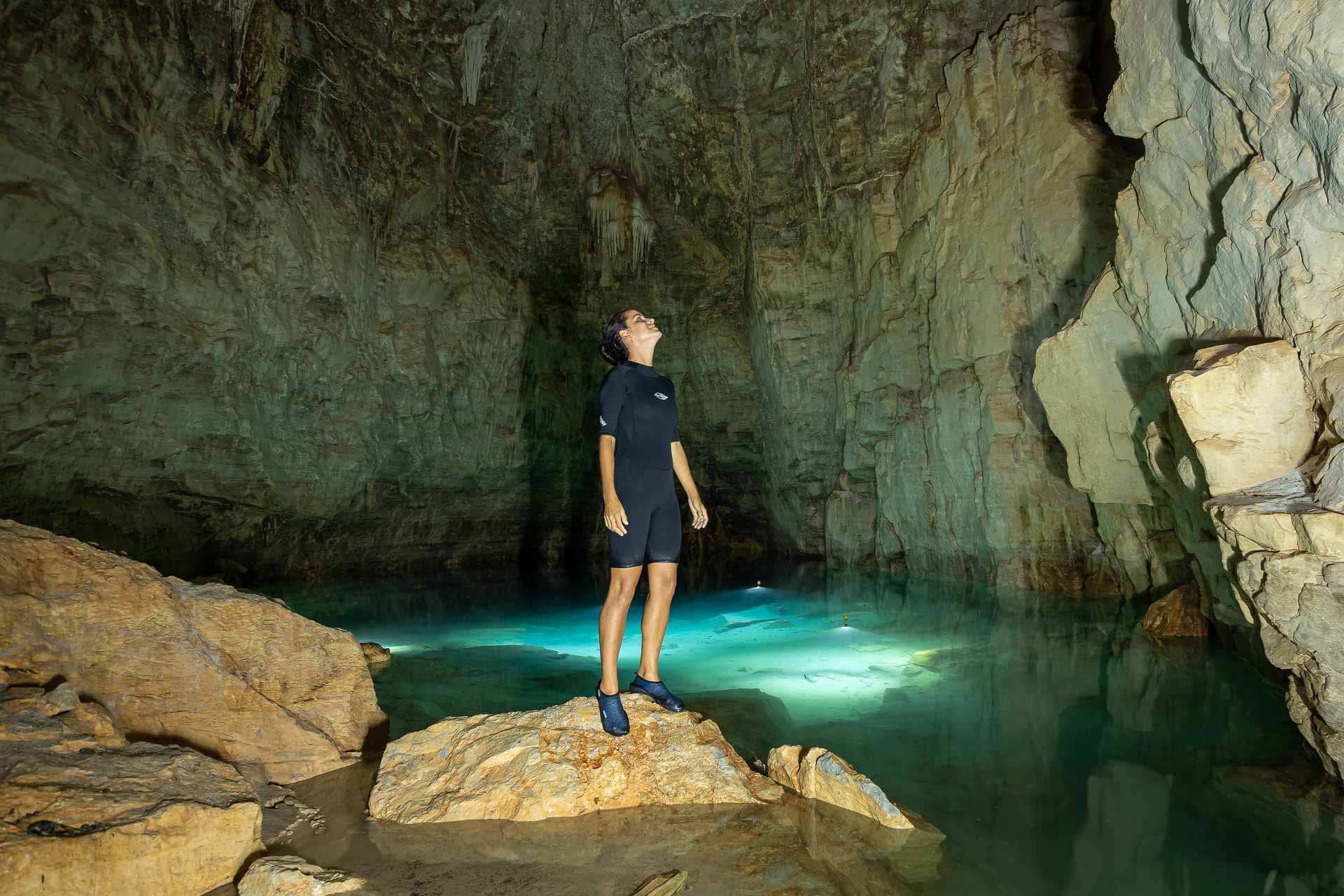 Fernanda in the middle and on top of a rock inside a cave with crystal blue water behind