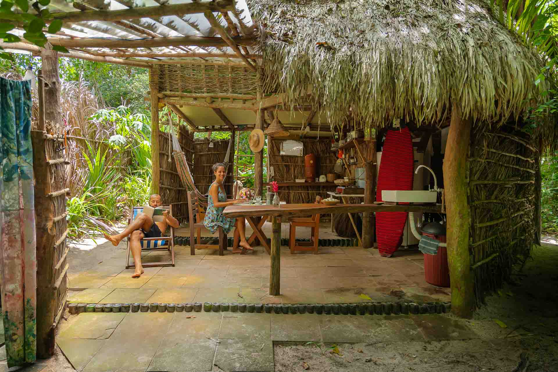 a bungalow in morere made of wood and straw with tiago and fernanda sitting down near a surf board