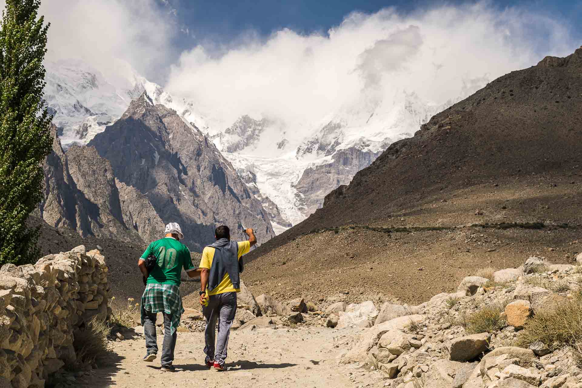 Tiago and a Pakistani walking in dirty road in between mountains with a snowed peak mountain in the back