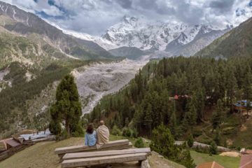 Tiago e Fernanda sentados no banco de madeira olhando para a montanha de Nanga Parbat coberta de neve na vila de Fairy Meadows, rodeada de pinheiros