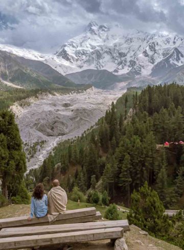 Tiago and Fernanda sitting on a bench looking at Nanga Parbat snowed peak mountain surrounded by a forest in Fairy Meadows