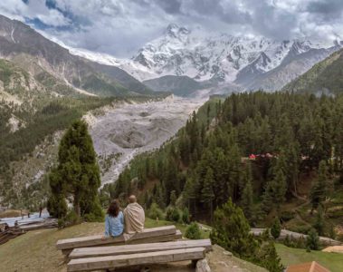 Tiago and Fernanda sitting on a bench looking at Nanga Parbat snowed peak mountain surrounded by a forest in Fairy Meadows