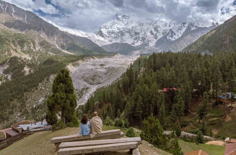Tiago and Fernanda sitting on a bench looking at Nanga Parbat snowed peak mountain surrounded by a forest in Fairy Meadows