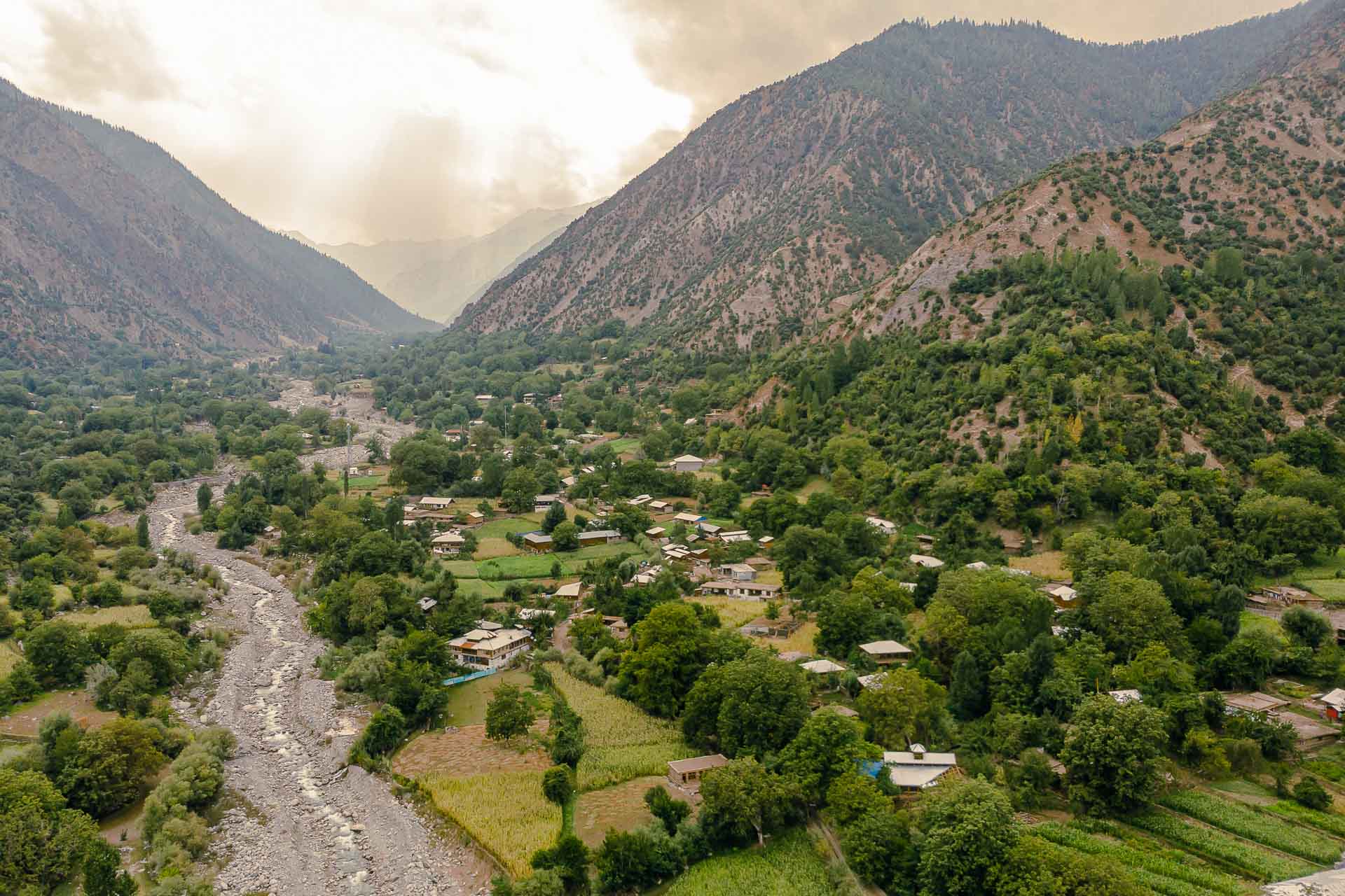 Vista aérea do Vale de Kalash com o sol no meio das nuvens e um rio seco ao lado da vila e casas