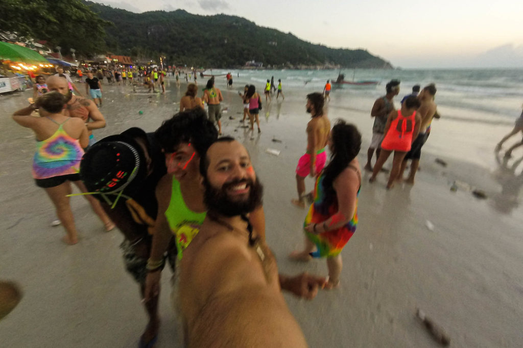 Tiago fazendo uma selfie no meio da praia de manhã em volta das pessoas dançando até o sol nascer durante a festa da Lua Cheia