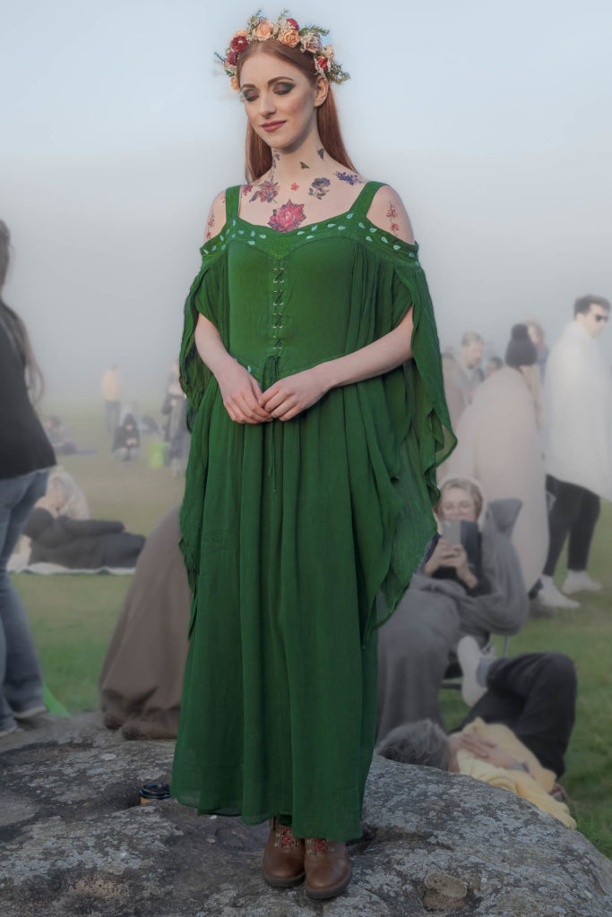 Mulher vestida de verde com tatuagens pelo corpo posando para a foto, com olhos fechados e segurando suas mãos, com a cabeça baixa
