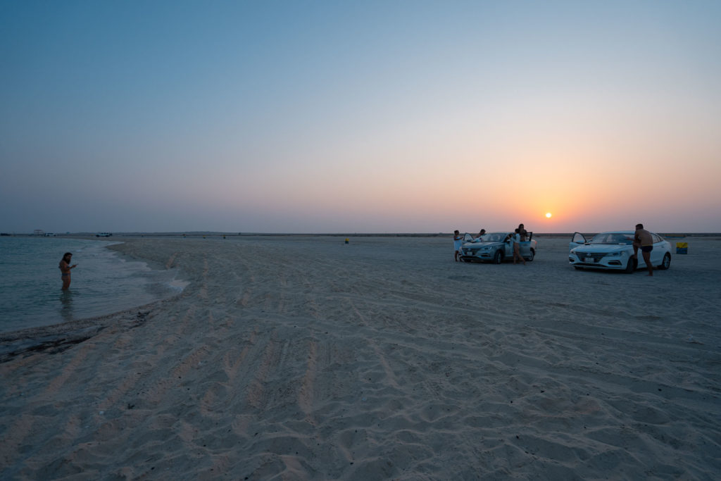 Uma das praias do Catar com alguns carros parados na areia e o sol de pondo ao fundo