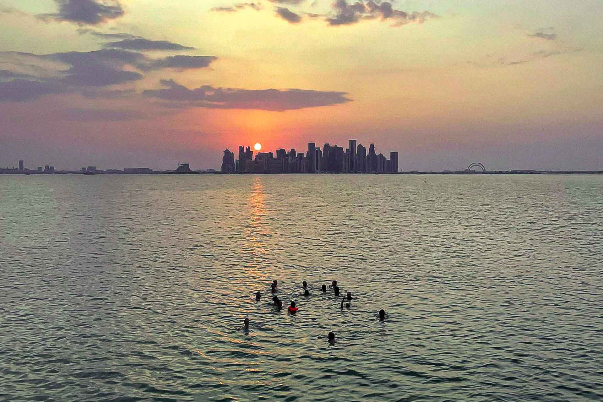 Vista panorâmica do mar da cidade de Doha com um grupo de pessoas na agua e o sol se pondo atras dos prédios altos da cidade