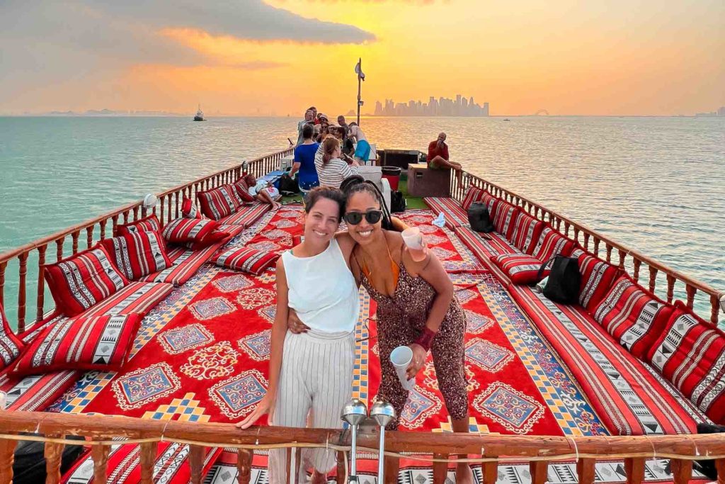 Fernanda hugging a friend on top of a boat in the sea of Qatar with the buildings in the background and a colourful sky