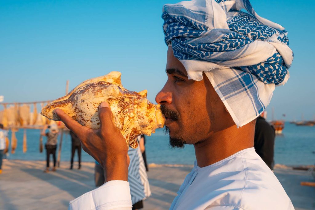 Um homem segurando uma grande concha na boca a usando de flauta com um turbante na cabeça e vestido tradicionalmente