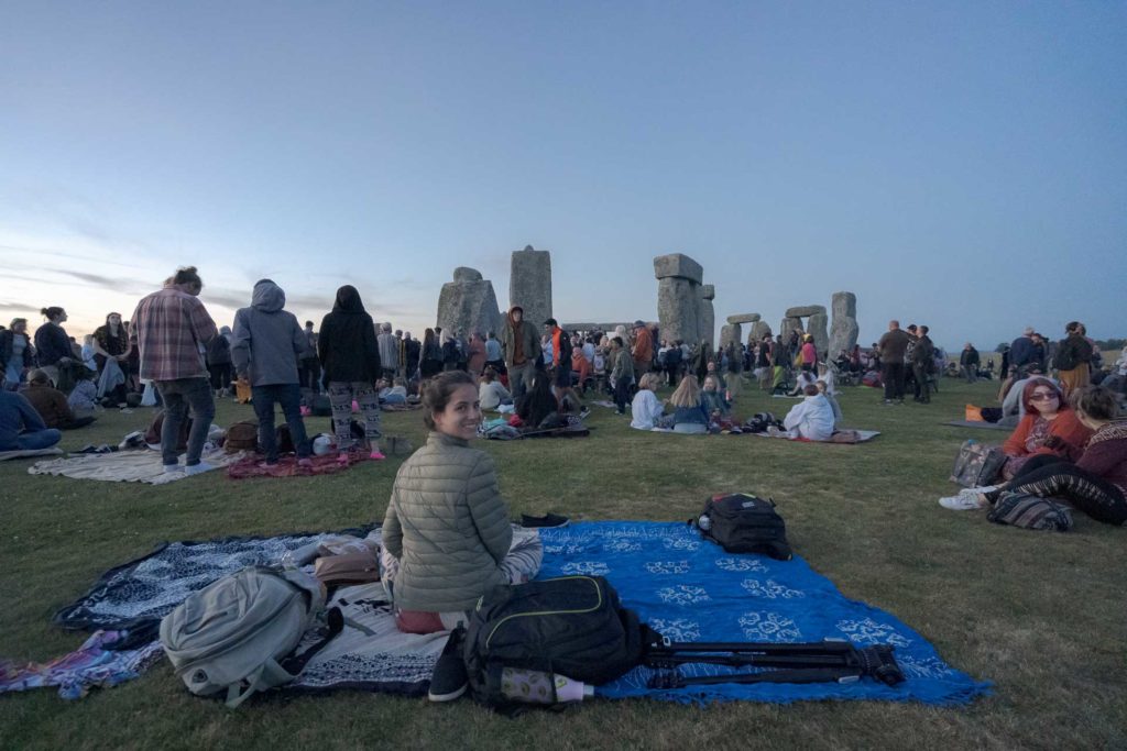 Fernanda sentada em cima de uma canga e olhando para a camera como as pedras de Stonehenge ao fundo durante o solstício de verão