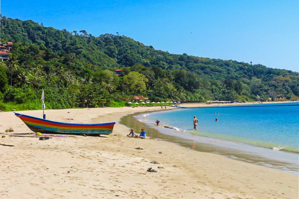 A white sand beach with a colourful boat parked in the sand with a few people bathing in the water