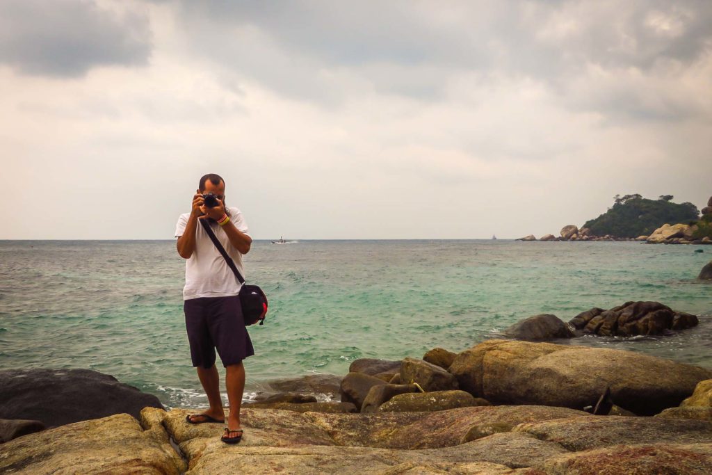 Tiago segurando a camera como se fosse tirar uma foto olhando para a lente e com o mar ao fundo